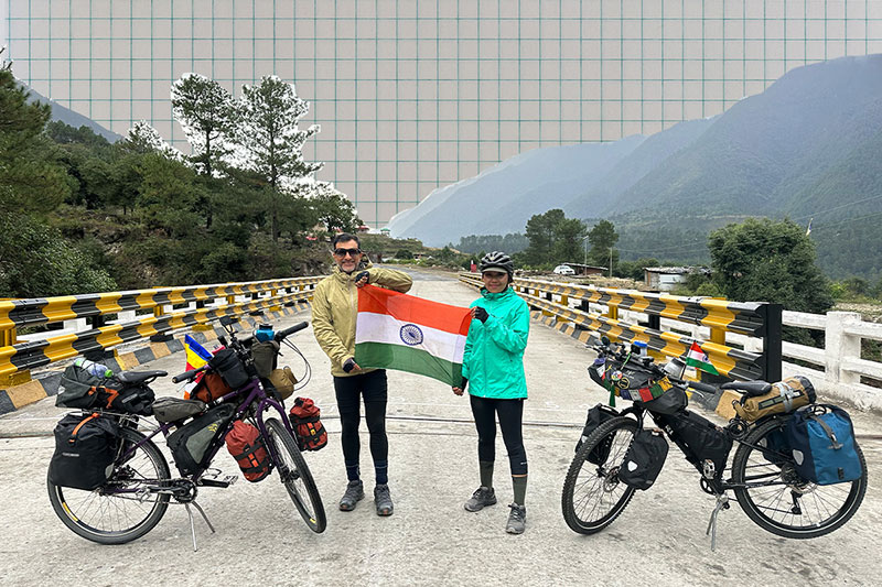 Dhruv and Ashim standing together holding flag on mountain road with loaded touring bikes
