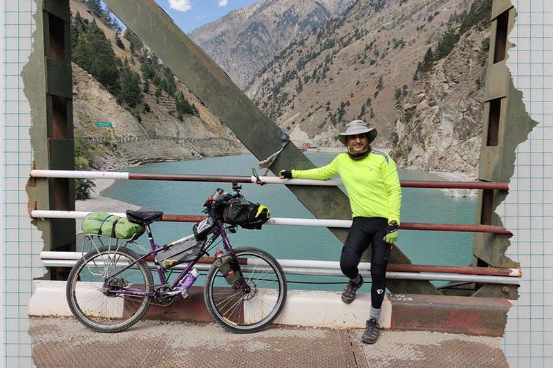 Dhruv standing with loaded bike on bridge with river and mountains in background