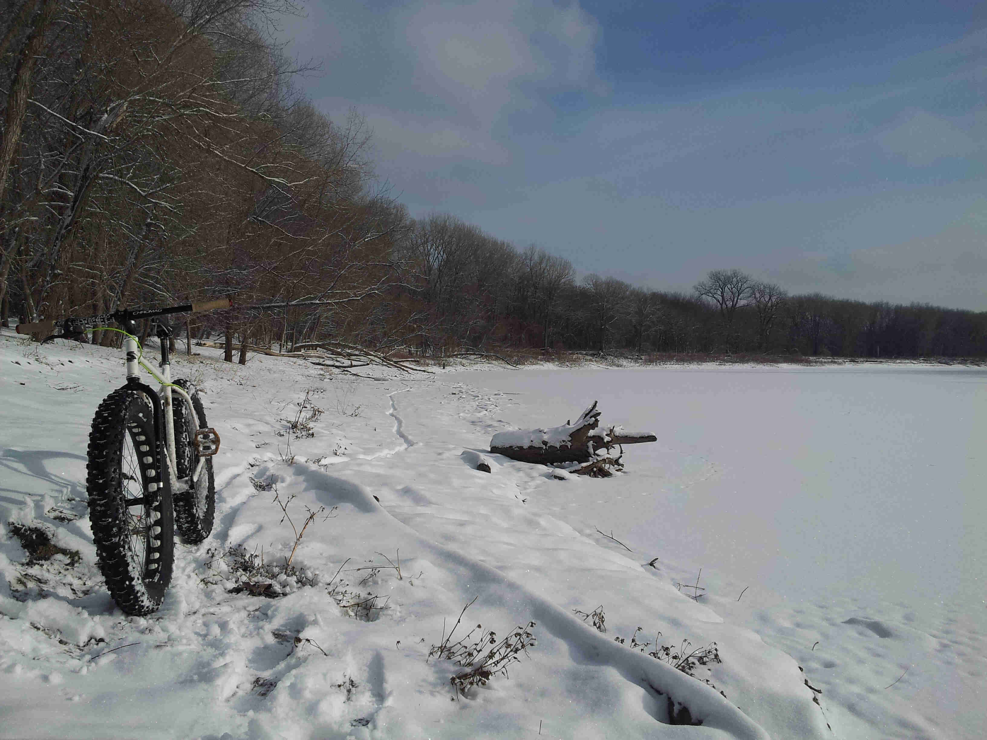 Front view of a white Surly Pugsley bike, on a snowy covered trail, beside a frozen river with bare trees on the sides