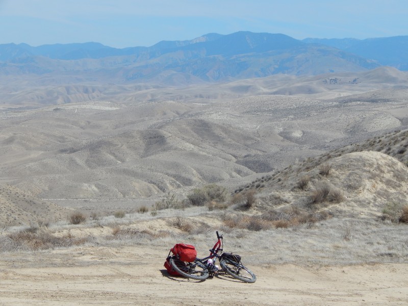 Right side view of a Surly bike with gear packs, laying on a dirt road, above desert hills and mountains in background