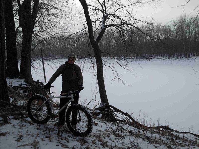Right side view of a white Surly Pugsley fat bike with a cyclist behind, on top of a bank with a frozen river below