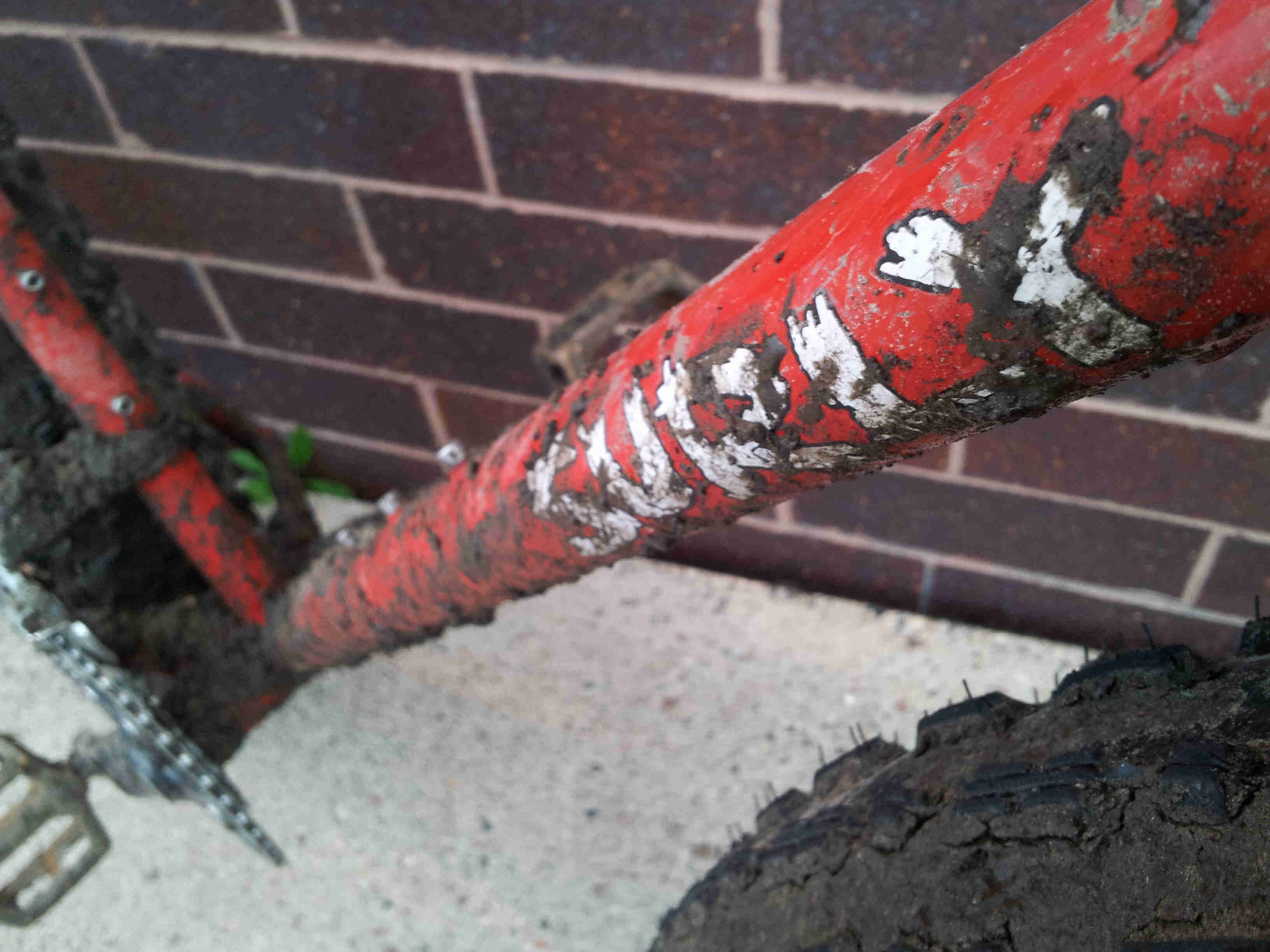 Close up, right side view of the muddy downtube of a red Surly Moonlander fat bike, with a brick wall behind it
