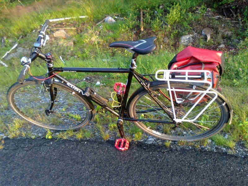 Left side view of a black Surly Cross Check bike, parked in grass & gravel, next to pavement