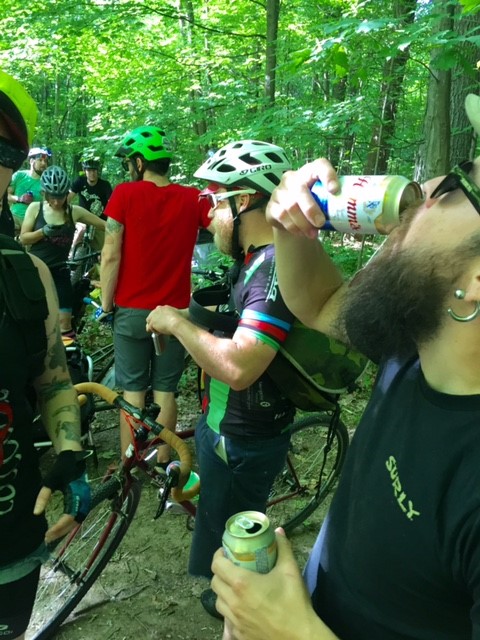 A group of cyclists, standing in the woods, with the one in the forefront, drinking a can of beer
