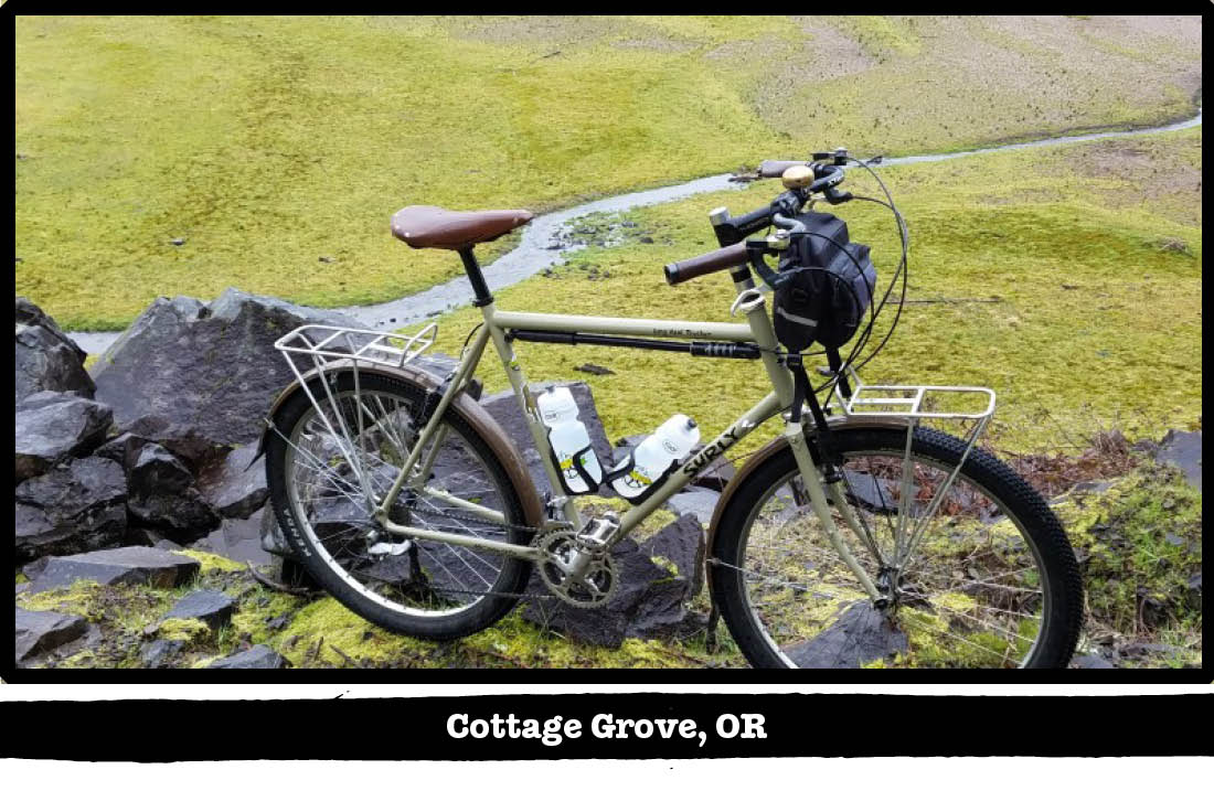 Right side view of a Surly bike on a cliff ledge, with a grass field and river below - Cottage Grove, OR tag below image
