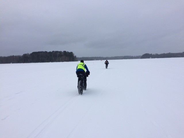 Rear view of 2 cyclists riding their fat bikes across a frozen, snow covered lake