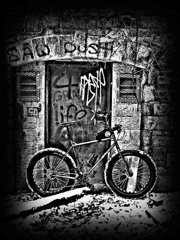 Black and white image - right side view of a Surly fat bike, parked on snow, in front of a door and wall with graffiti