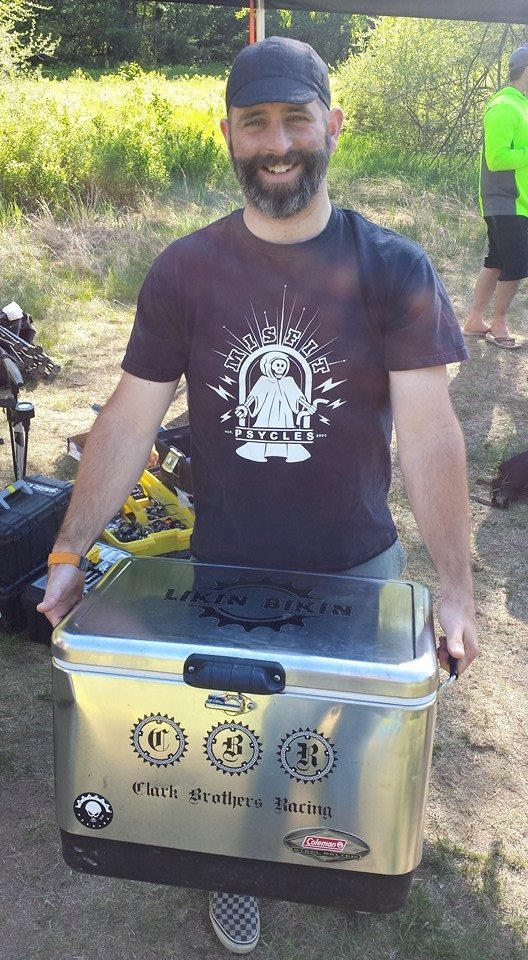 Front view of a person posing, wearing a Misfit Cycles t-shirt, holding a silver cooler in front of them