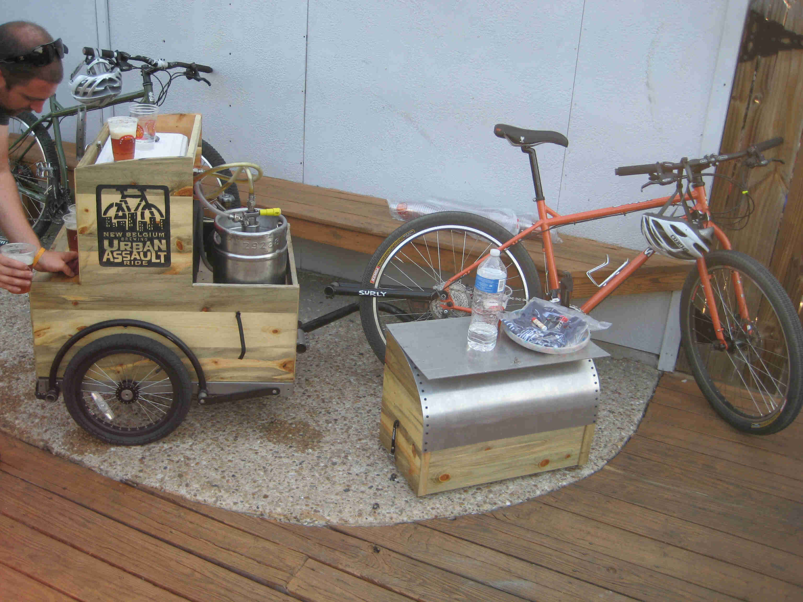 Right side view of an orange Surly bike & trailer with keg inside, on an outside deck, with a person behind the trailer