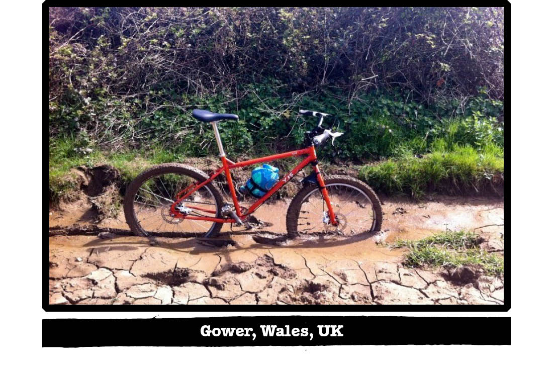 Right side view of a red Surly bike with it's tires buried in a muddy trail - Gower, Wales, UK tag below image