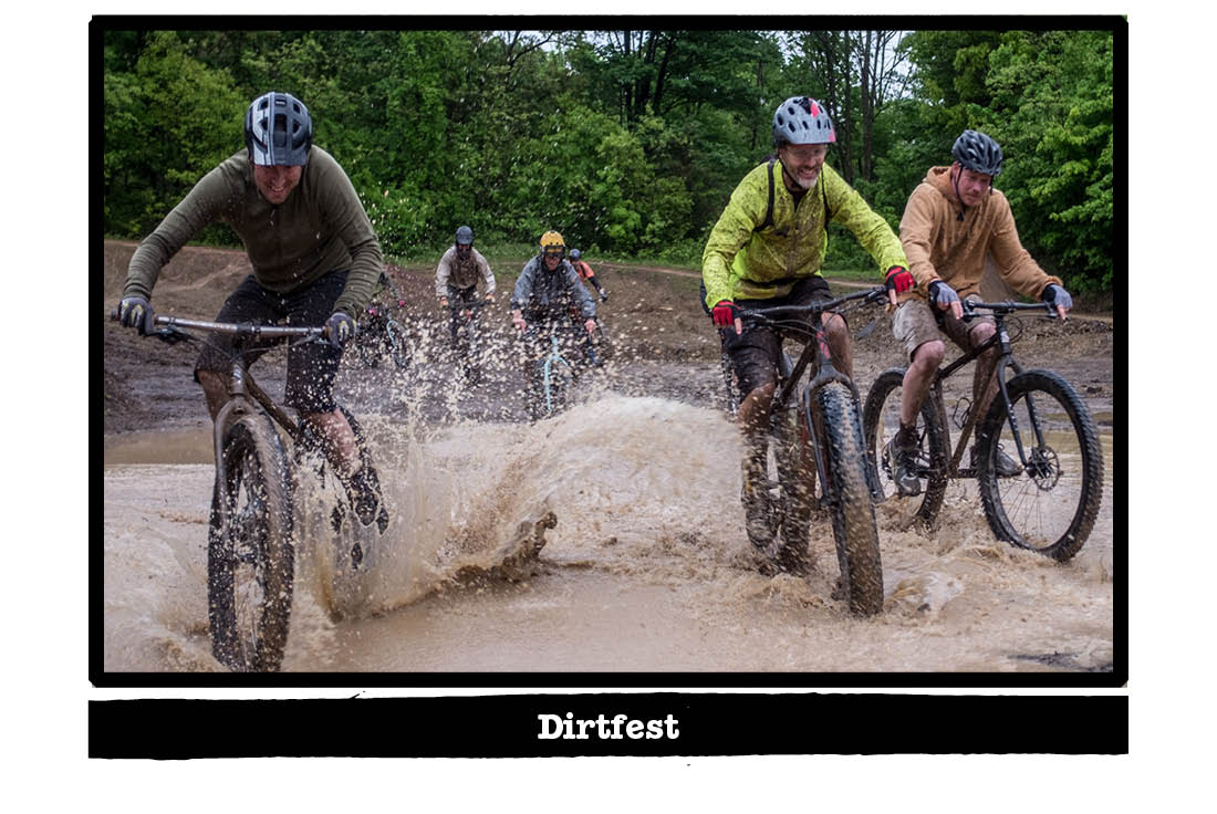 Front view of cyclists riding through mud puddle, with trees in the background - Dirtfest tag below image