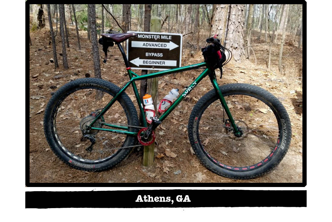 Right profile of a Surly bike, green, leaning on a sign post, on a dirt trail in the woods - Athens, GA tag below image