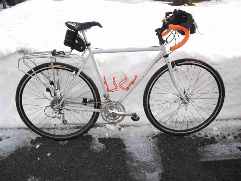 Right side view of a white Surly road bike, on an icy, paved trail, alongside a tall snow bank