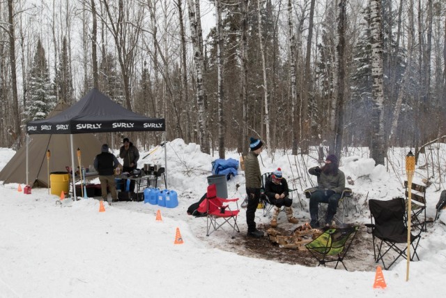 People gather around a snowy campsite with a Surly canopy, and trees in the background