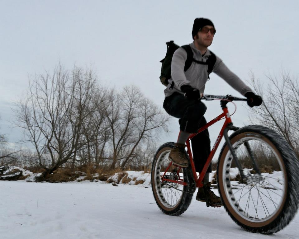 Upward, front right side view of a cyclist riding a red Surly fat bike on snowy ground, with bare trees in background