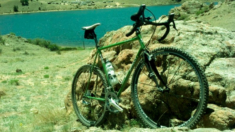 Front, right side view of a green Surly bike, facing up a hill, leaning on a rock, with a lake in the background