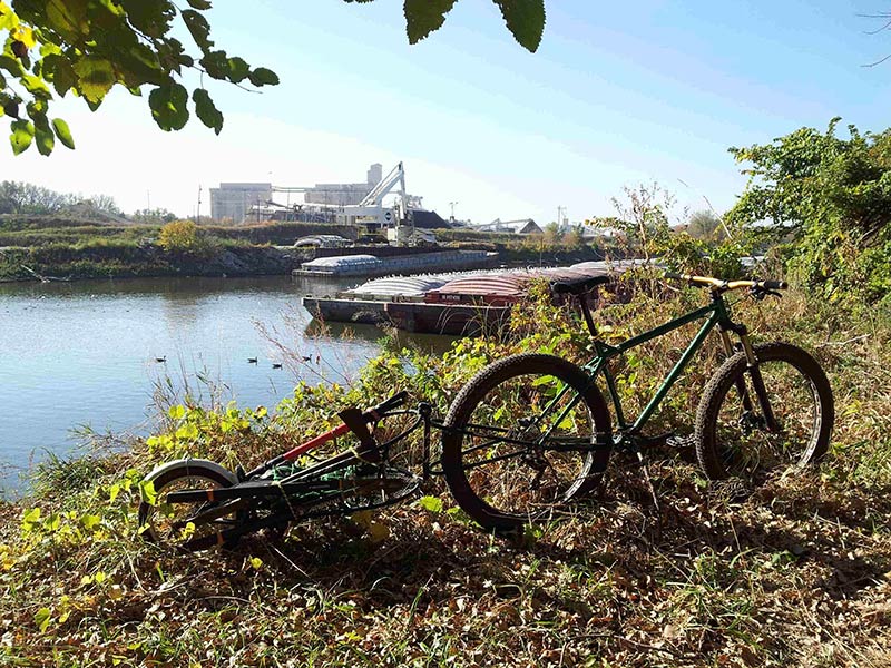Right side view of a green Surly Krampus bike and trailer, parked on a grassy bank, with a barge in a river behind it