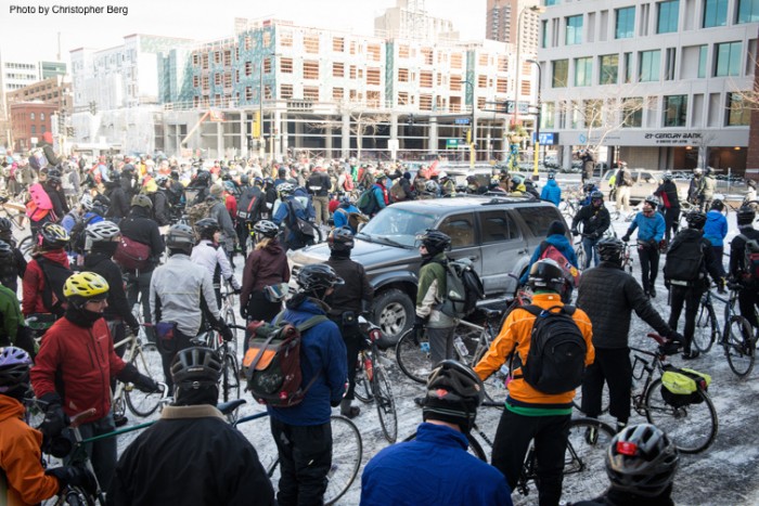 A large group of winter cyclists and their bikes, gather on a city block with buildings in the background