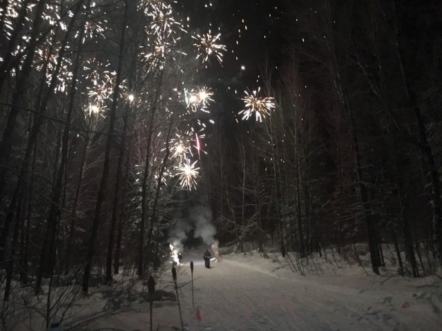 Front view of cyclist riding down a snowy trail with trees on the side, at night with fireworks exploding above