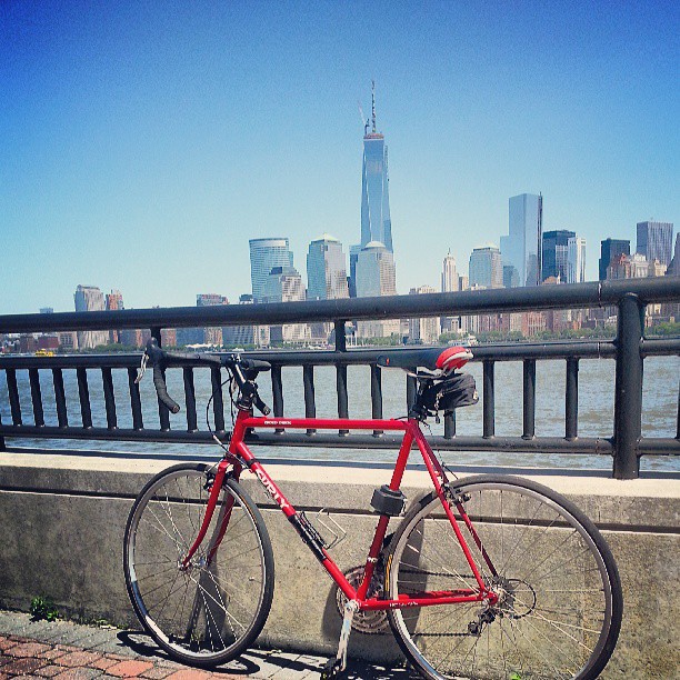 Left side view of a red Surly Cross Check bike, leaning on a cement wall with a railing, and a bay city in background