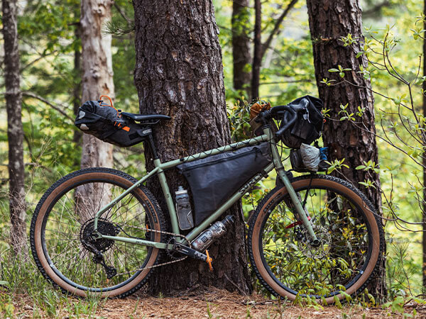 Ghost Grappler leaning against tree in forest with seat pack, framepack, handlebar bag, front rack, and two water bottles