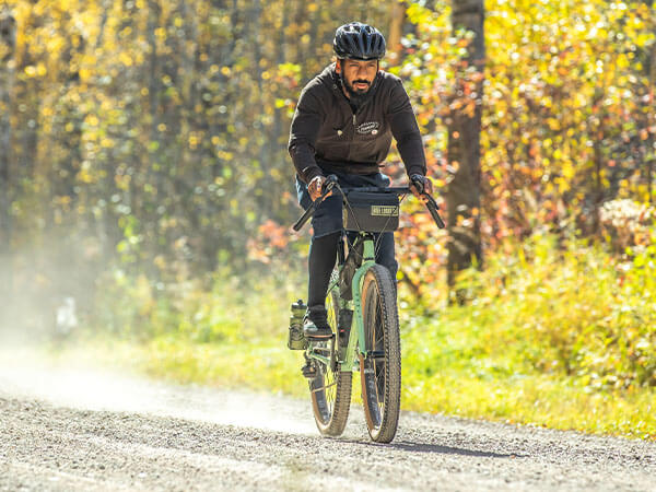 Person riding dusty gravel road on loaded Grappler on sunny day, thick forest in background