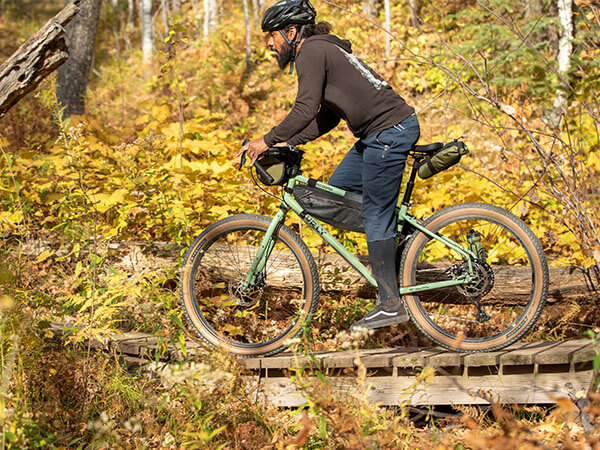Person riding Ghost Grappler bike with small frame pack, seat pack, and handlebar bag in forest across small wooden bridge