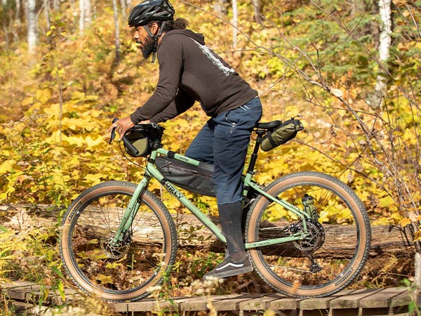 Person riding Grappler bike with small frame pack, seat pack, and handlebar bag in forest across small wooden bridge