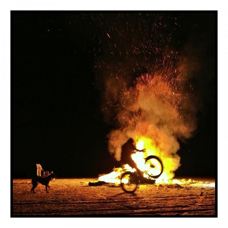 Right side silhouette of a cyclist popping a wheelie on a bike, in front of a bonfire at night 