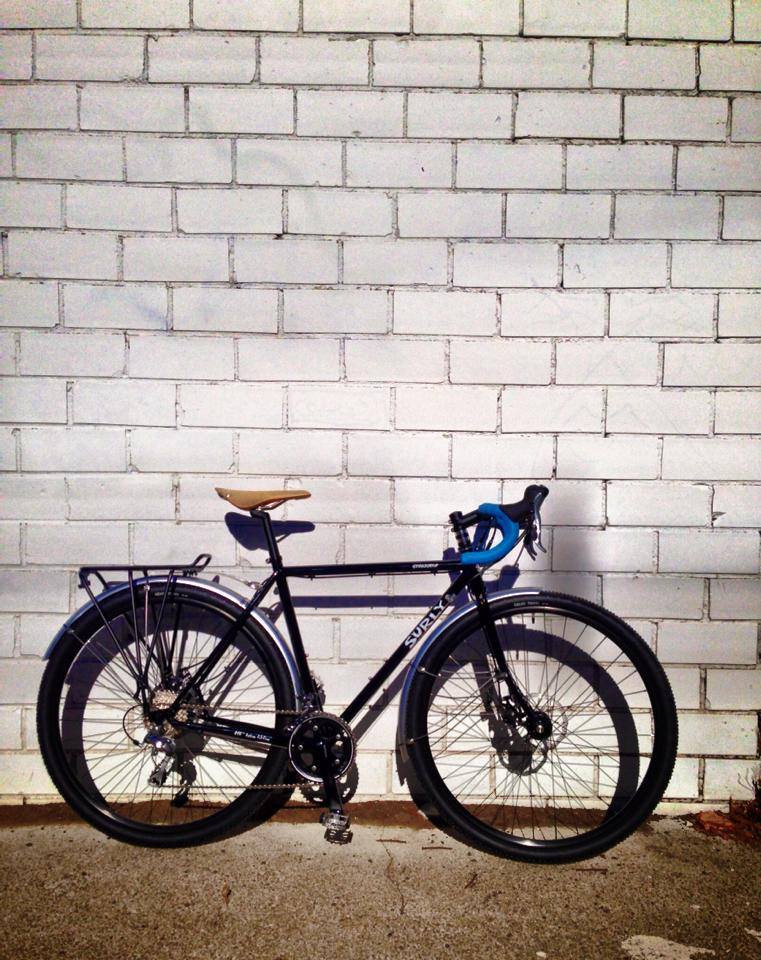 Right side view of a black Surly bike, parked on sand, leaning against a white brick wall