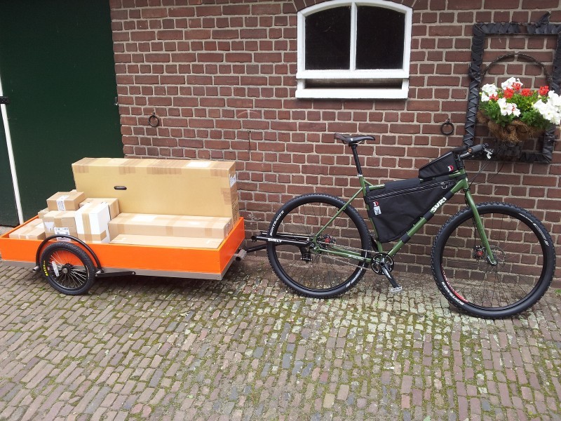 Right side view of a green Surly bike, with frame bag and a loaded trailer, leaning against a brick wall on a brick road