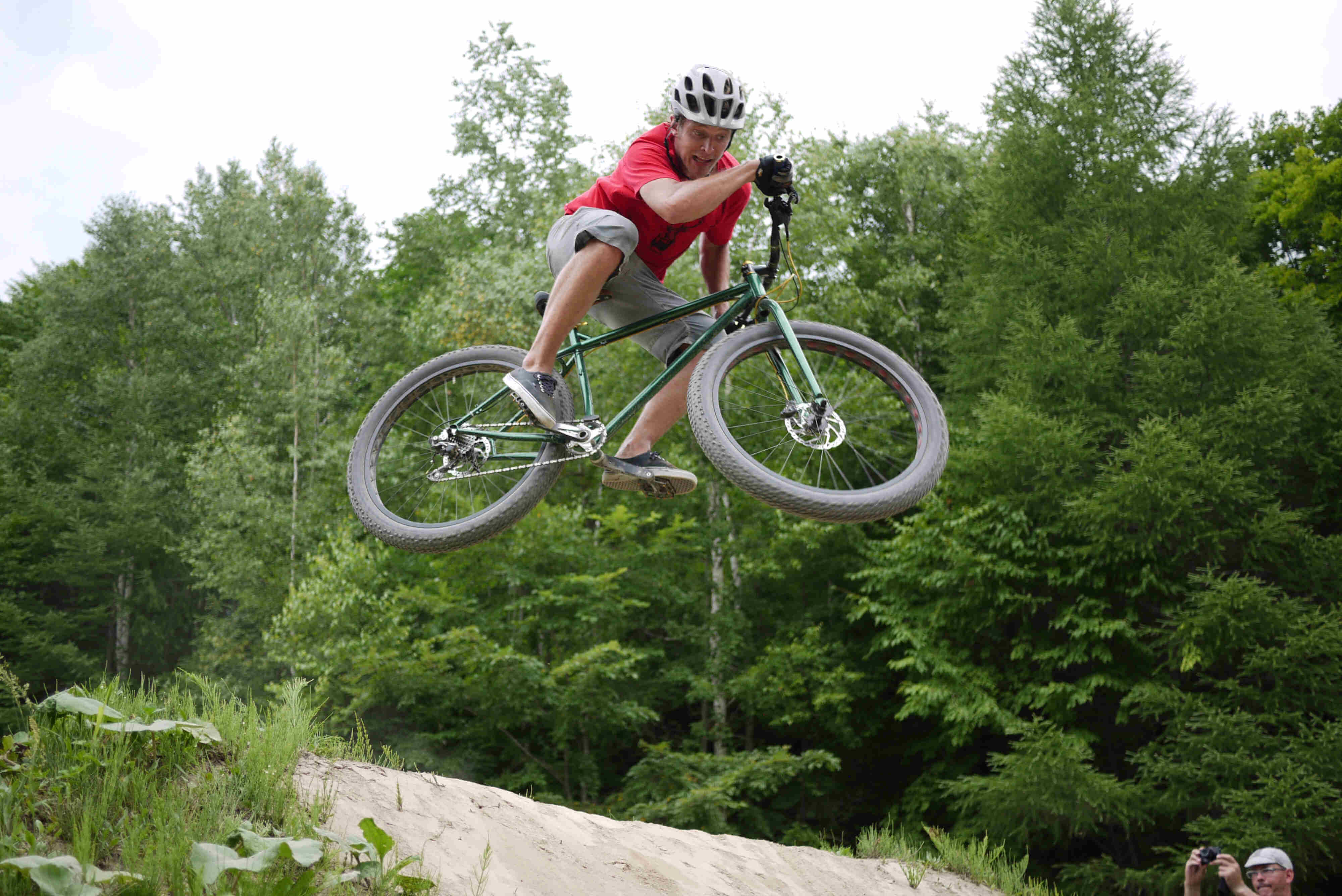 Right side view of a cyclist, on a green Surly Krampus bike, launching off of a dirt ramp with trees in the background