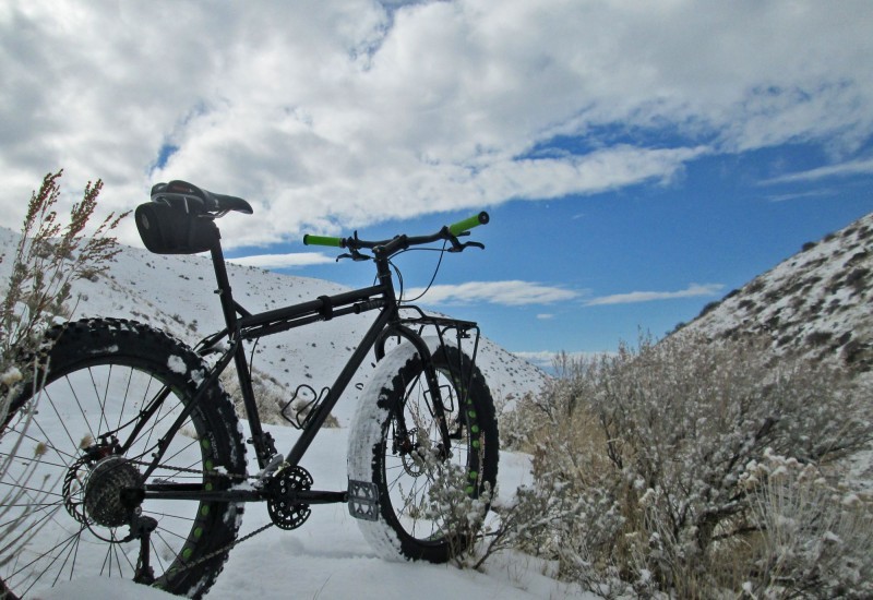 Rear, right side view of a black Surly fat bike, parked on snowy ground next to a bush, facing snow covered hills