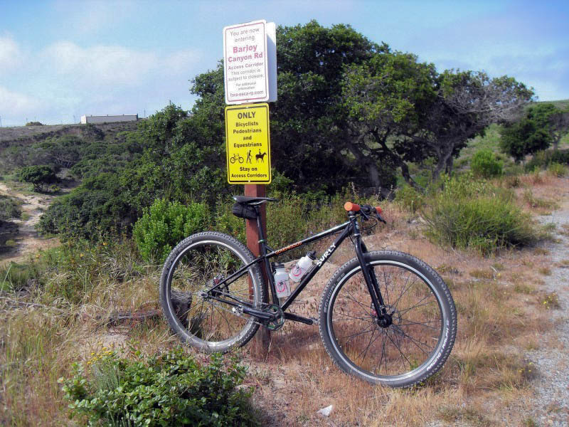 Right side view of a black Surly bike, parked against a park sign in a brushy field with trees in the background