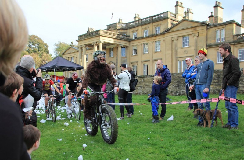 Front view of a cyclist, in a werewolf costume, riding a Surly fat bike on a grass course, with a large mansion behind