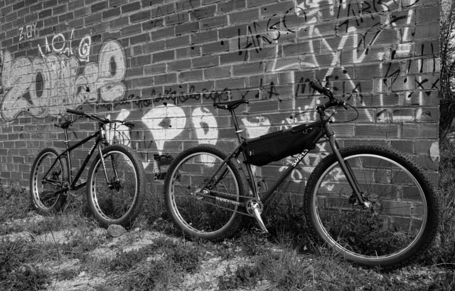 Right side view of 2 Surly bikes in single file, leaning against a brick wall with graffiti on it, 
