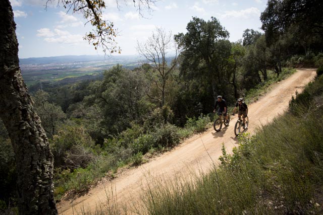 Front, left side view of 2 cyclists, riding their bikes down a gravel road along a hill, overlooking a valley of trees