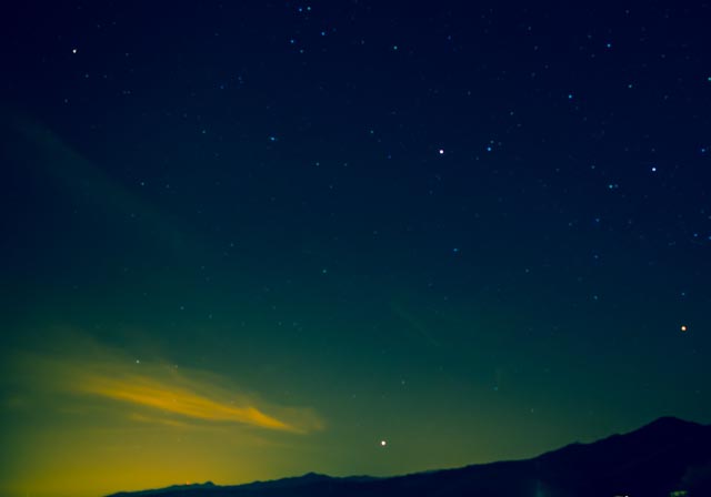 Nighttime view of the sky with a green glow and stars, above mountain hills