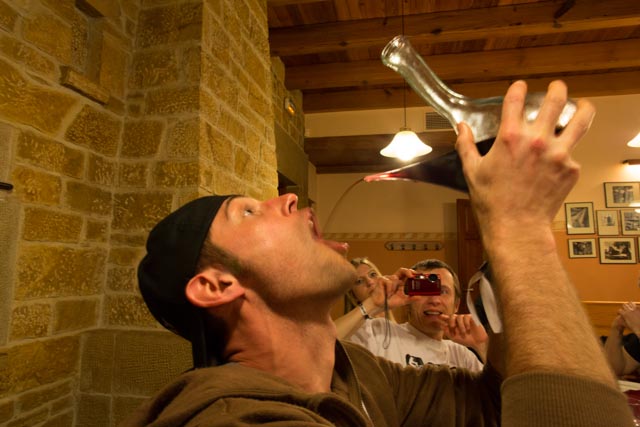Right side view of a person holding up and decanter, pouring wine into their mouth, inside a room with people watching
