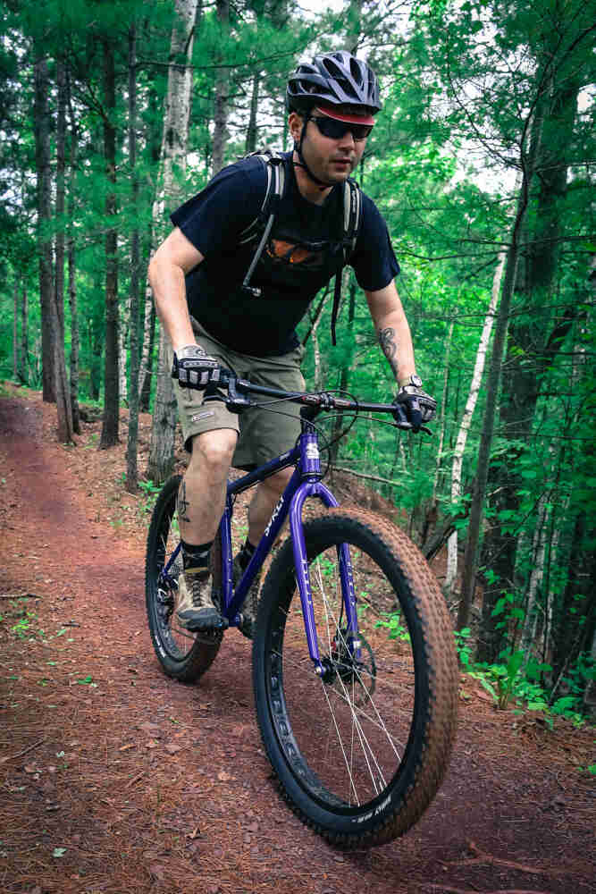 Front view of a cyclist riding a purple Surly Krampus bike, down a red dirt trail in a green forest
