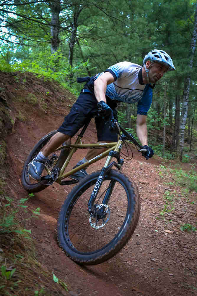 Right side view of a cyclist riding a Surly Instigator bike across a red dirt berm in the forest