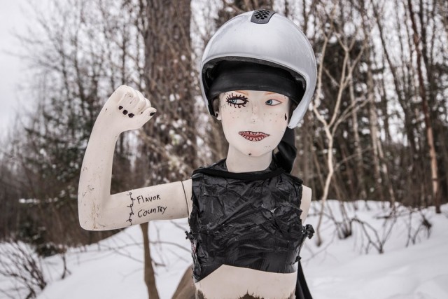 Front view of a mannequin with a helmet, holding an arm up, with snow and trees in the background 