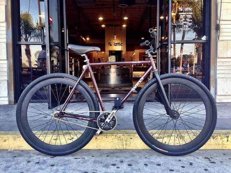 Right side view of a burgundy Surly bike, parked against a street curb, in front of a coffee shop