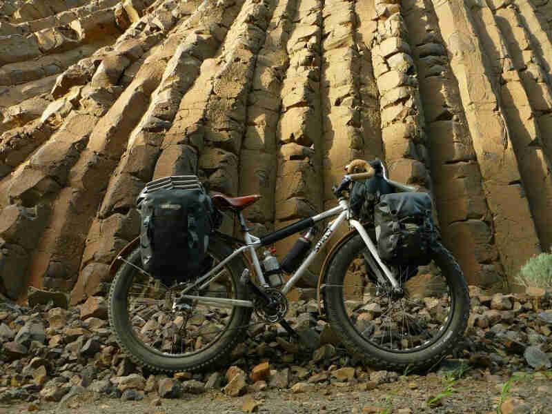 Right side view of a white Surly fat bike, loaded with gear, parked on rocks at the bottom of a stone cliff wall