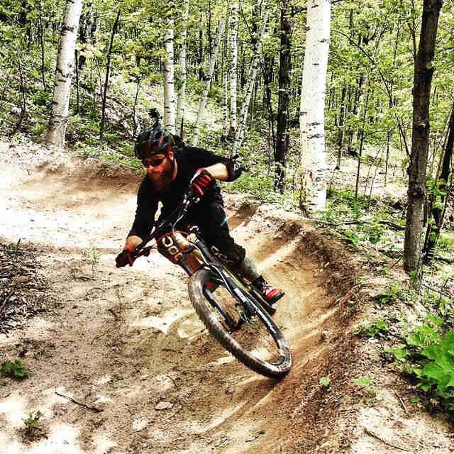 Front view of a cyclist, riding a Surly Instigator bike, rounding a berm on a dirt trail in a forest