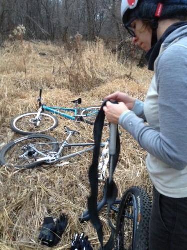 Left side view of a cyclist, standing in tall brown grass with their bike laying next to them, fixing a tire innertube