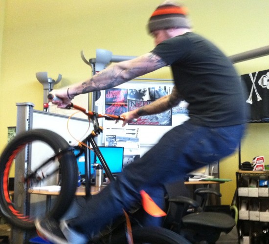 Left side view of a person in an office cubicle, popping a wheelie on a black Surly Pugsley fat bike