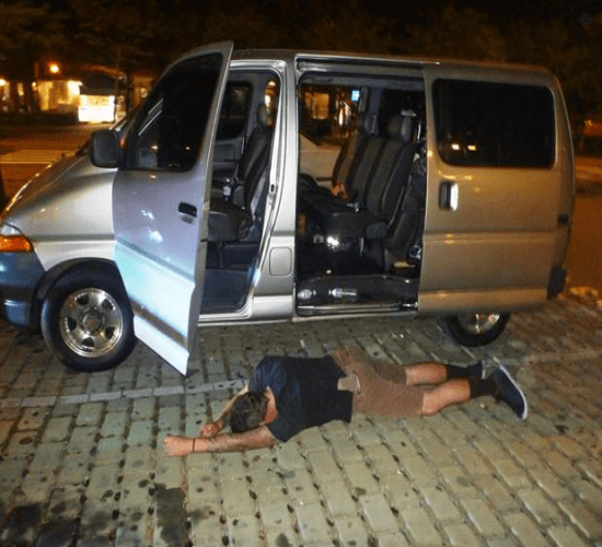 A person laying on a brick sidewalk at night, along the left side of a silver van with all it's doors open
