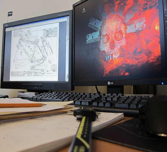 Front view of 2 computer monitors on a desk, with a CAD drawing of a bike on the left, and skeleton graphic on the right