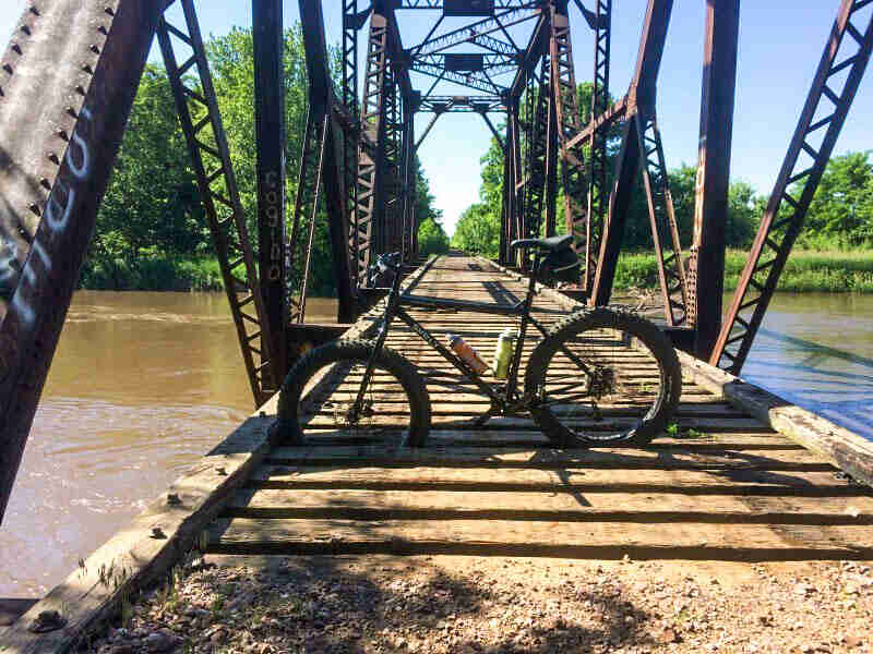 Left side view of a Surly fat bike, parked across a railroad tie decked bridge over a river